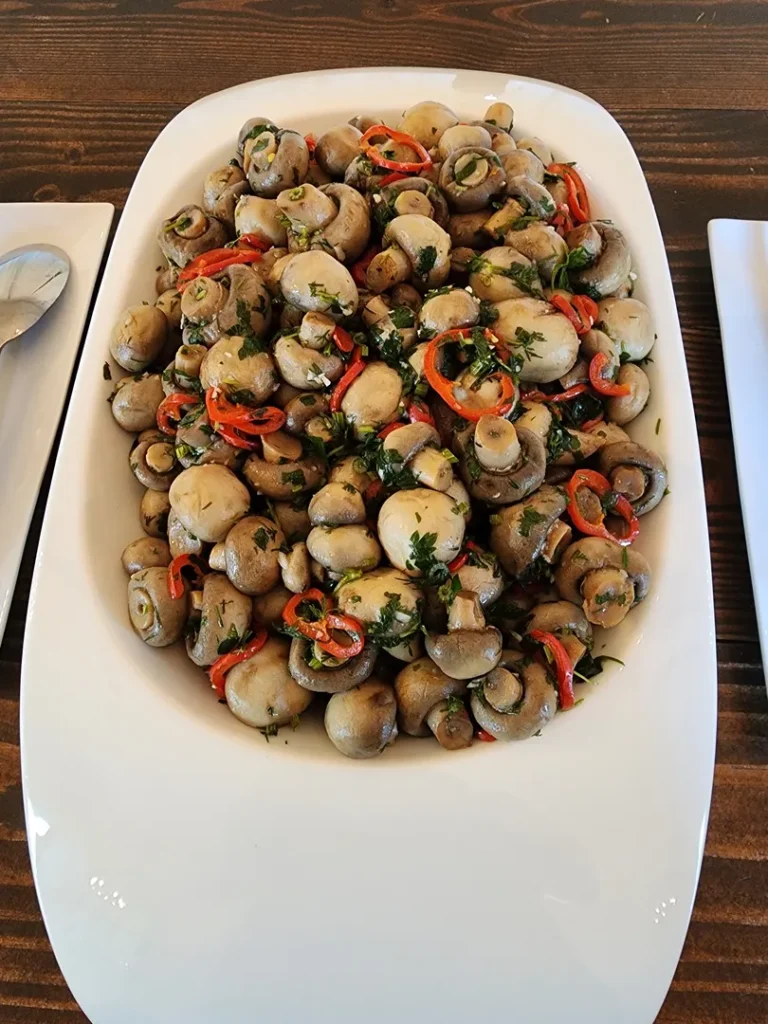 A white rectangular dish filled with marinated mushrooms garnished with sliced red chili and chopped herbs, set on a wooden table.