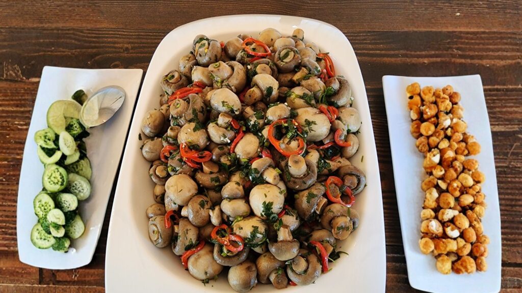Three white serving dishes display sliced cucumbers with lime, a salad of whole mushrooms with herbs and red chili, and a snack of roasted chickpeas on a wooden table.