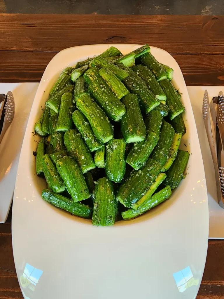 A large white platter filled with chopped cucumbers coated in herbs or seasoning, set on a wooden table next to metal serving tongs.