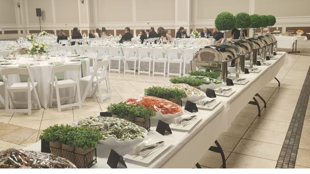 A long buffet table with covered trays and salads is set up in front of round dining tables with guests seated at an indoor event venue.