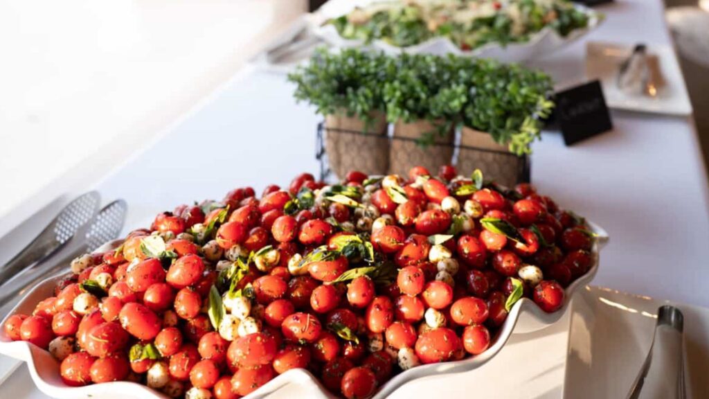 A large white serving dish filled with cherry tomatoes, mozzarella balls, and basil sits on a buffet table next to green potted plants.