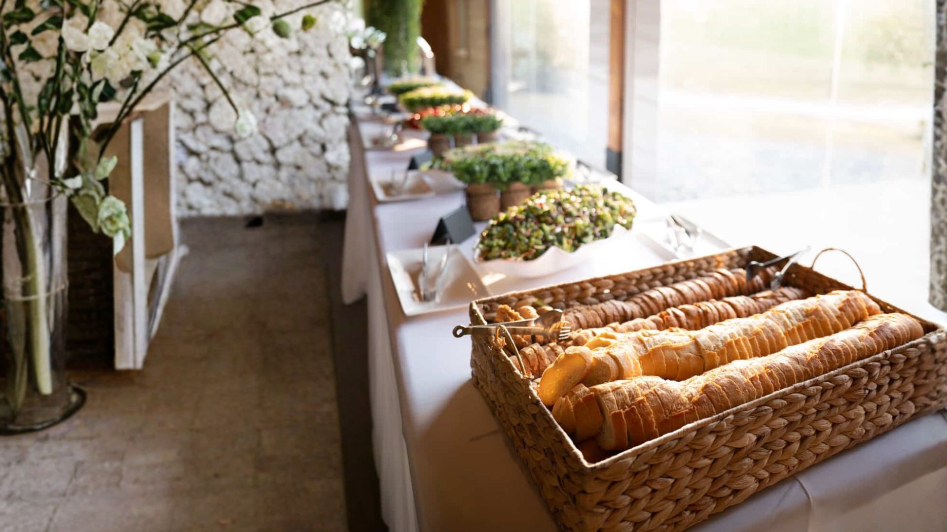 A buffet table with assorted bread in a wicker basket in the foreground and various salads and dishes arranged in a row in the background.