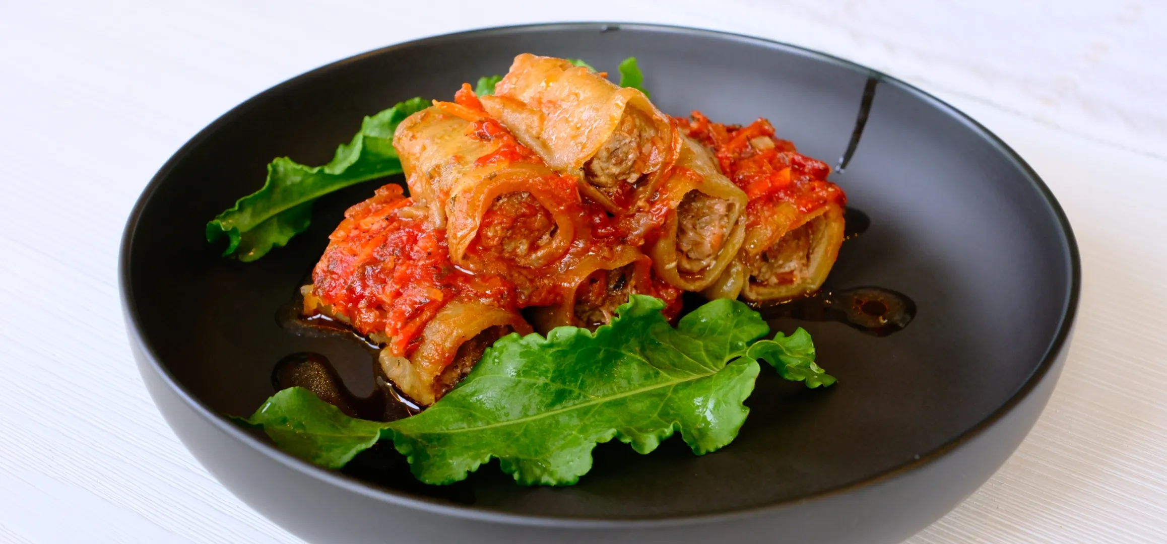 A black plate with stuffed cabbage rolls in tomato sauce, garnished with green leafy vegetables.