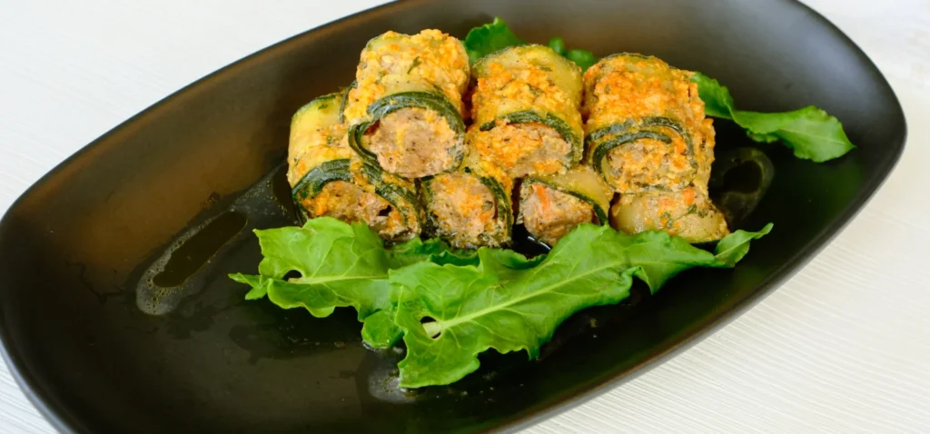 Rolled zucchini stuffed with filling and baked, served on a black plate with leafy greens as garnish.
