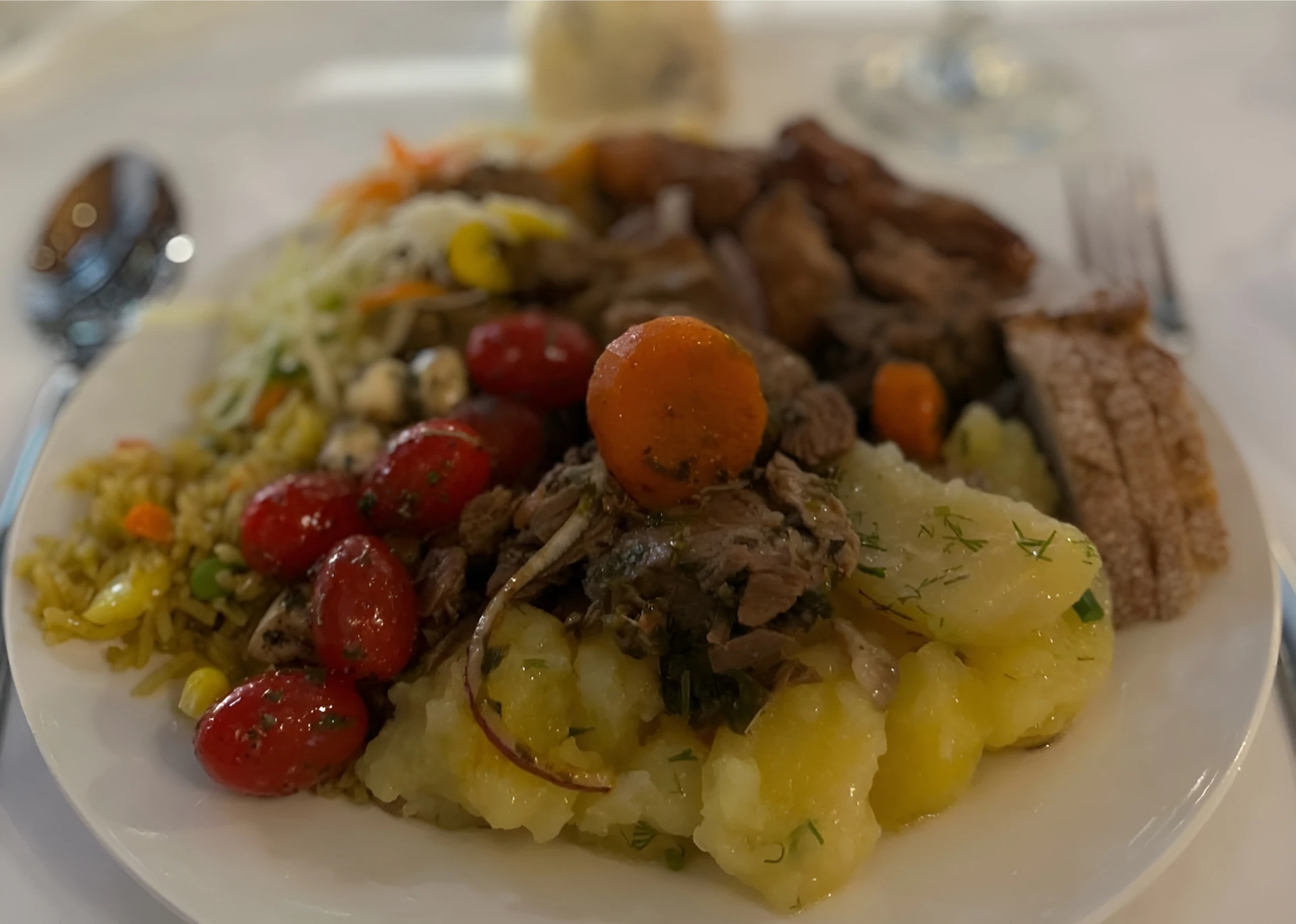 A plate of food featuring cooked potatoes, cherry tomatoes, shredded meat, bread, salad, and grains, with a spoon and fork nearby.