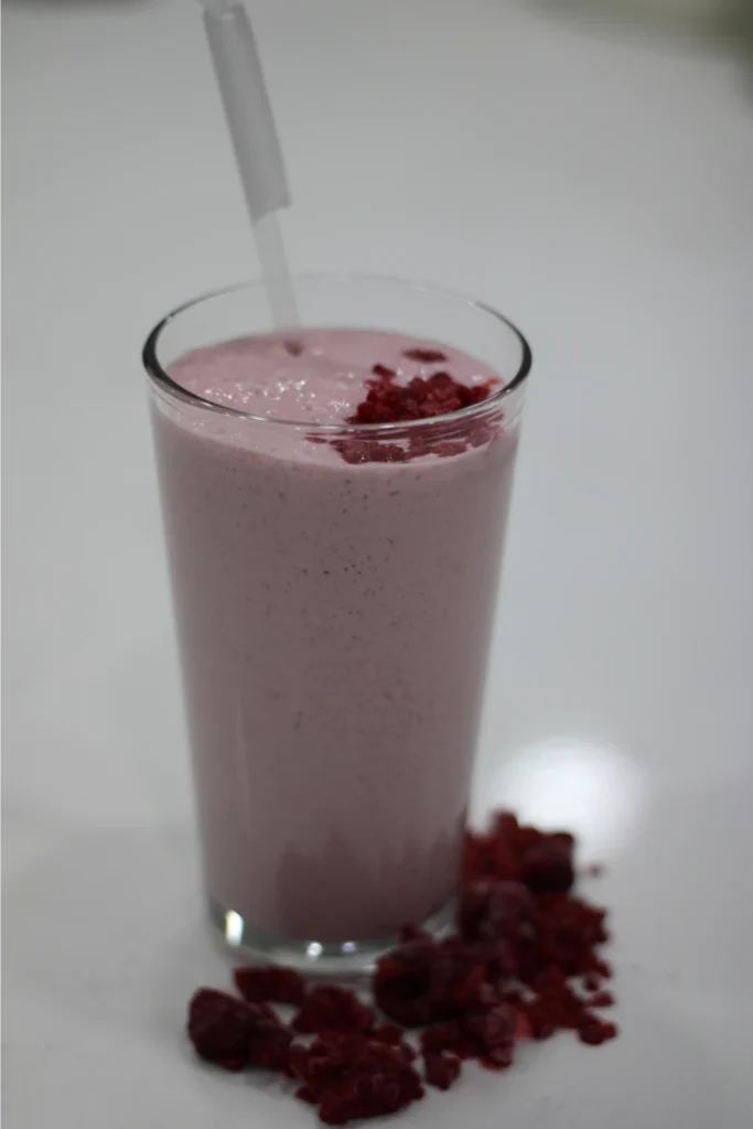 A tall glass of pink smoothie with a straw, topped with small pieces of dried fruit. Some dried fruit pieces are scattered beside the glass on a white surface.