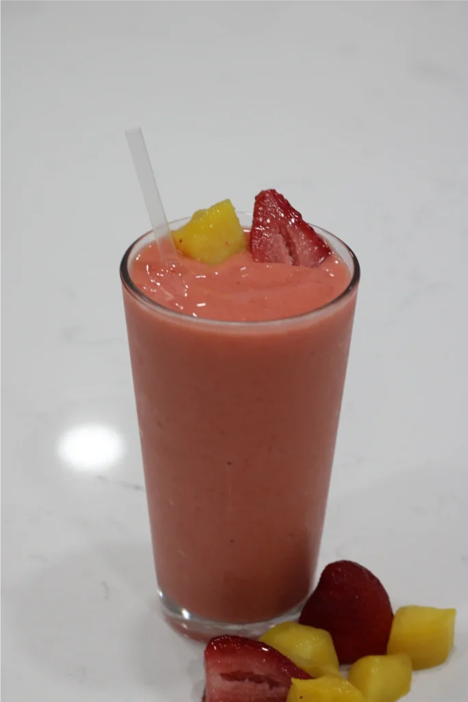 A glass of pink smoothie with a straw, garnished with pieces of strawberry and pineapple, placed on a white surface.